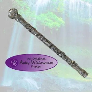 Wooden wand with ornate metal spherical top and textured bark surface against a misty forest waterfall background