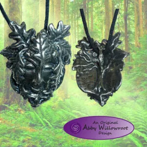 Two black metal pendants shaped like stylized green man faces with leaf designs, shown front and back views against a forest background