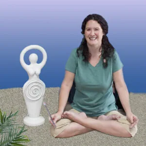 Woman in green shirt sitting cross-legged next to white spiral goddess statue on sand with blue background