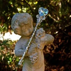 Stone cherub figurine holding a decorative wand with heart-shaped crystal top, surrounded by evergreen foliage
