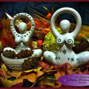 Two white ceramic goddess figurines with moon-shaped heads and decorative patterns, one holding a bowl with pinecones, surrounded by autumn leaves and dried flowers