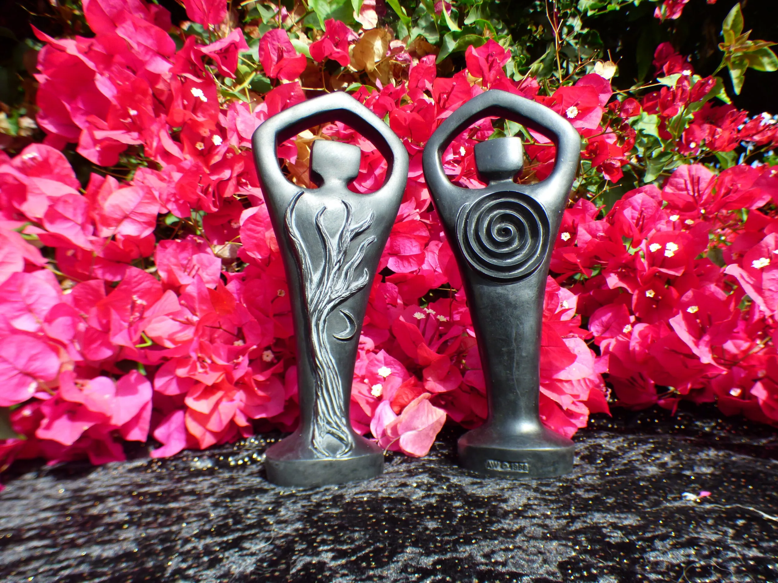 Two decorative metal goddess figurines with ornamental designs displayed against vibrant pink flowering shrubs in a garden setting.
