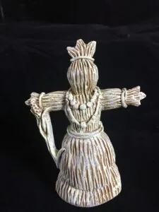 Whitewashed wooden corn dolly figurine with outstretched arms holding a bundle, tied at waist and neck against dark background