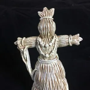 Whitewashed wooden corn dolly figurine with outstretched arms holding a bundle, tied at waist and neck against dark background