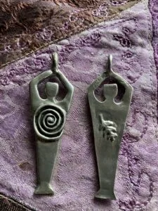 Two pewter pendants with human-form bodies and raised arms, left featuring a spiral symbol, right featuring a hand symbol, on purple textured fabric