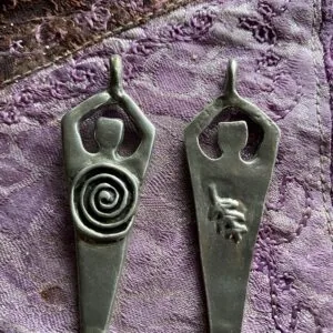 Two pewter pendants with human-form bodies and raised arms, left featuring a spiral symbol, right featuring a hand symbol, on purple textured fabric